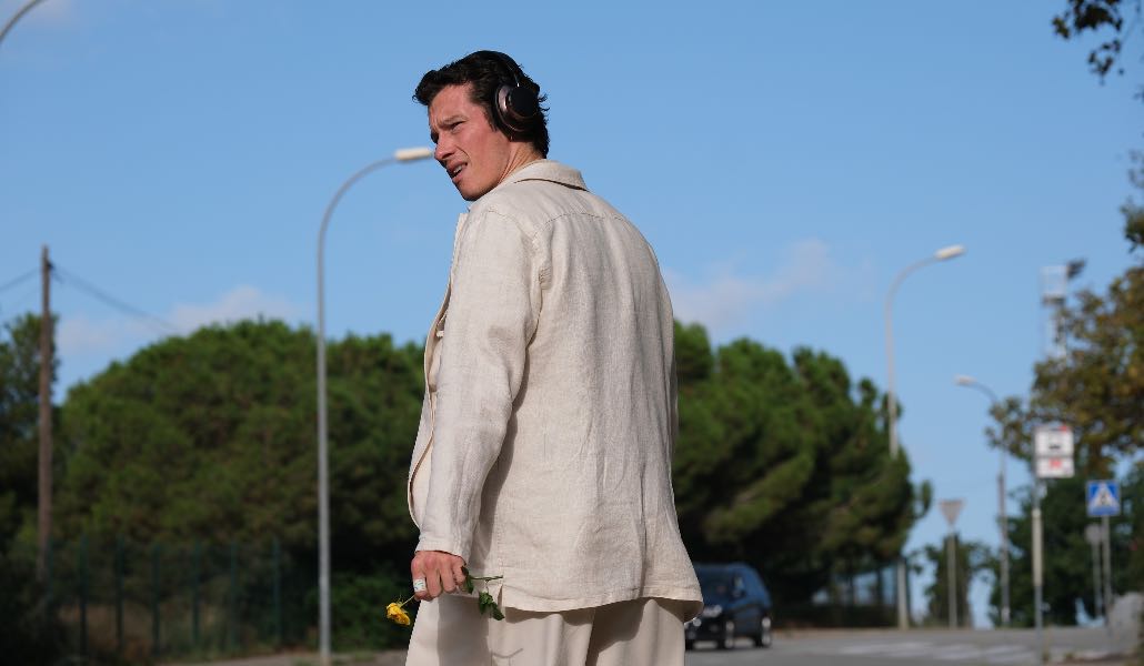 Callum Turner in una scena del film 'Rosebush Pruning', che cammina lungo una strada girato indietro, indossando un abito chiaro, con le cuffie alle orecchie e una rosa gialla in mano